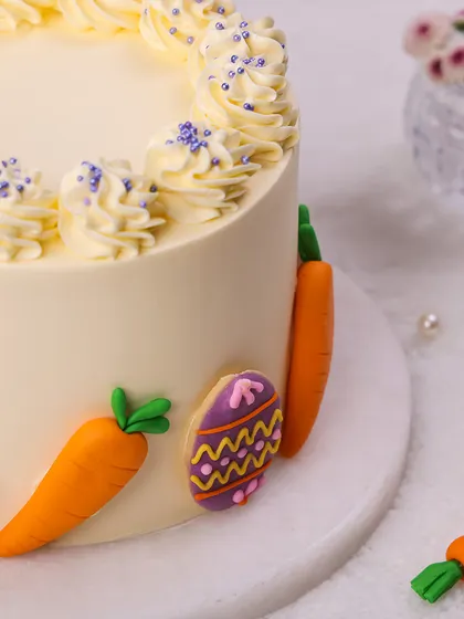 We decorate for the holiday with this festive Easter cake. A smooth, cream-colored cake is adorned with colorful, hand-decorated Easter egg cookies and fondant carrots, creating a bright and cheerful design.
