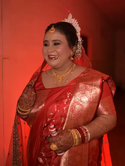 A candid photograph of a Bengali bride sharing a laugh. Dressed in a traditional red saree and holding a 'gach kouto', this image captures a moment of pure, unscripted joy.