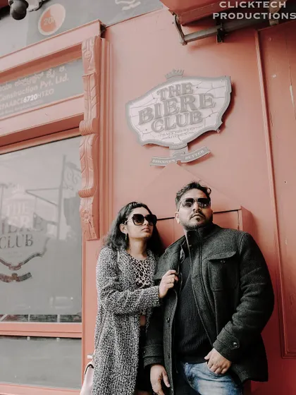A low-angle shot of a couple in stylish coats, posing against the wall of 'The Biere Club', creating a cool, urban portrait.