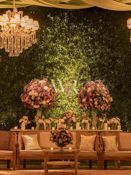 A beautiful lounge setup against a wall of lush greenery. The natural wood seating is complemented by large, colorful floral arrangements, creating a space that feels like an elegant garden room.