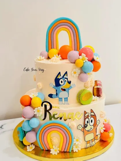 A colorful two-tier cake featuring the popular characters from Bluey, complete with rainbows and macarons.