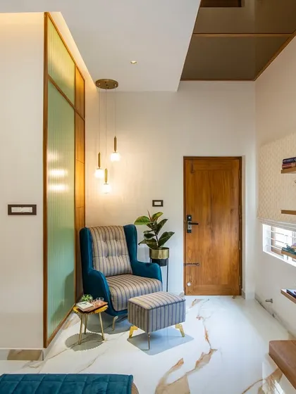 A cozy reading corner in a master bedroom, featuring a plush blue armchair and ottoman. The corner is defined by a frosted glass and wood partition, with warm pendant lights creating an intimate ambiance.