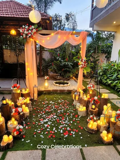 A rustic and romantic garden setup perfect for an anniversary or proposal. A wooden arch draped in peach fabric is surrounded by dozens of LED candles on tree stumps, with a path of scattered flower petals.