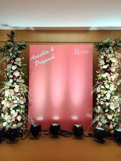 A clean and contemporary photo backdrop designed for a modern couple. The minimalist pink panel with personalized names is flanked by two towering pillars of white and blush florals, offering a chic and stylish spot for pictures.