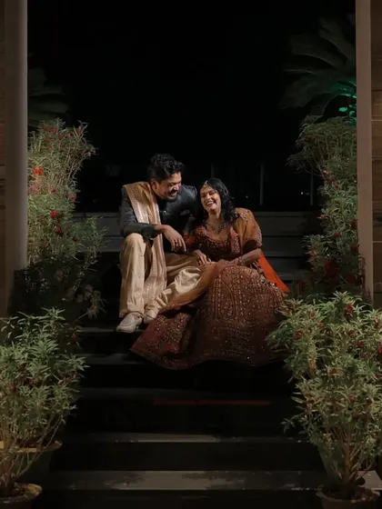 A couple shares a laugh while sitting on a staircase at night, surrounded by plants. This relaxed, candid portrait from their engagement shoot captures their easy and joyful chemistry.