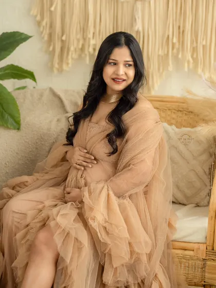 A soft and dreamy portrait of a mom-to-be in a ruffled beige tulle gown, seated on a cane sofa in our natural-light boho studio.
