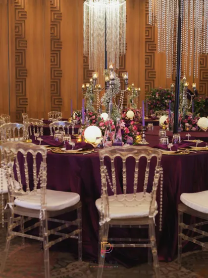 A tablescape that is pure drama. A deep purple tablecloth provides a rich base for gold charger plates, glowing orbs, and a towering candelabra centerpiece, all under the sparkle of a crystal chandelier.