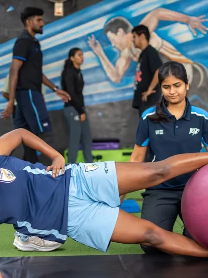 A female athlete performs a side plank with leg raise on a stability ball, guided by a physiotherapist. This exercise targets core and glute strength, crucial for injury prevention.