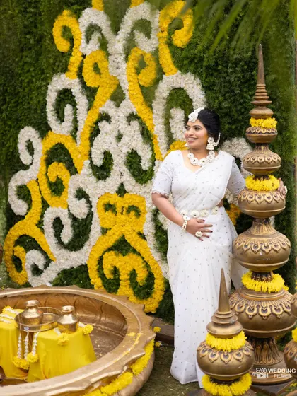 The bride poses next to a stunning floral wall of white and yellow, her white saree complementing the elegant Haldi decor.