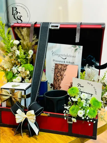 A housewarming or anniversary gift presented in a stylish red and black trunk. It's filled with fresh green and white flowers, a personalized card, and other home-centric gifts to celebrate a new beginning.