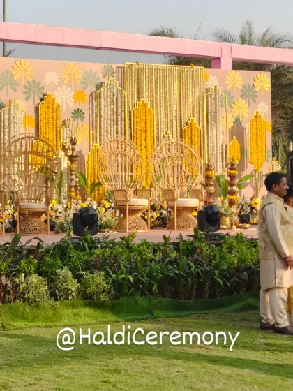 A stunning Haldi ceremony stage with a backdrop of floral patterns and hanging marigold strings. The ornate golden chairs add a touch of royalty to the vibrant setup.
