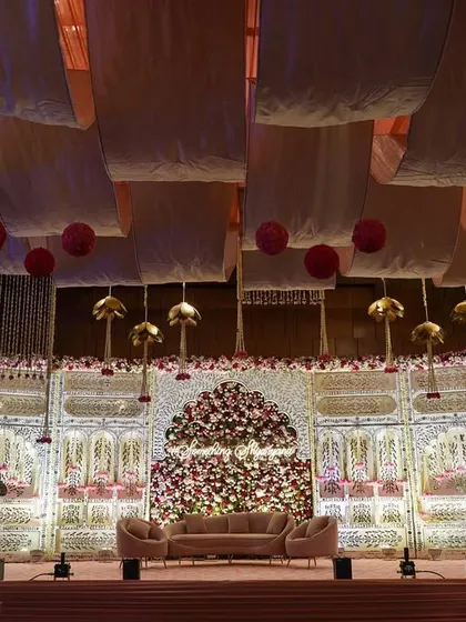 A stunning stage setup combining traditional jali work with a massive floral installation. The ceiling is draped with soft fabric panels and accented with hanging lotus-bud-shaped elements.