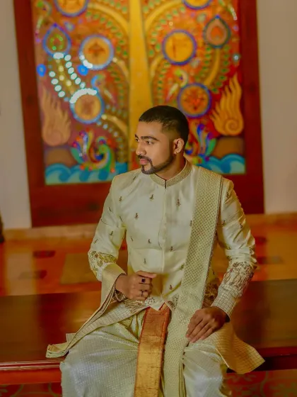 A seated portrait that offers a closer look at the fabric and embroidery detail of the off-white sherwani jacket.