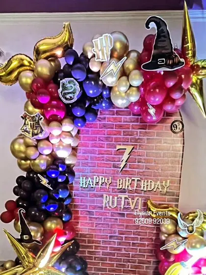 A detailed Harry Potter-themed backdrop for Rutvi's 7th birthday. The design includes a brick wall print, house logos, a sorting hat cutout, and a balloon garland in house colors, creating a magical photo corner.