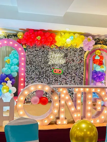 A colorful Cocomelon first birthday party. The stage is decorated with a silver shimmer wall, lighted arches, and large "ONE" marquee letters.