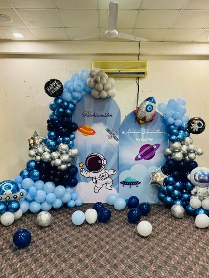 An out-of-this-world space theme party with custom astronaut and planet backdrops. The scene is framed by a galaxy-inspired balloon arrangement in shades of blue, silver, and navy.