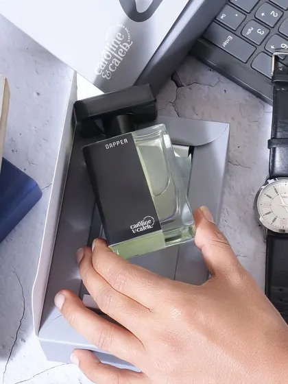 A lifestyle shot showing a hand taking the 'Dapper' perfume out of its box on a work desk. This action oriented shot makes the product feel like a personal, everyday essential.