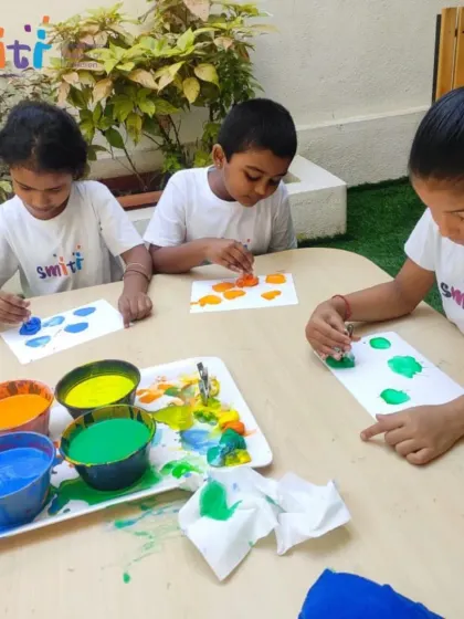 Children sit at an outdoor table, fully engaged in painting. Celebrating Holi through art allows them to experience the joy of the festival in a child-friendly way.