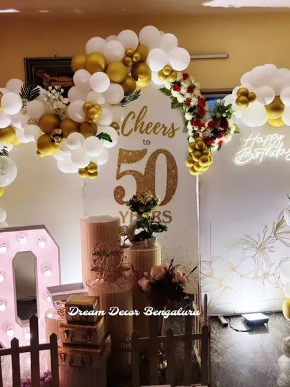 A 'Cheers to 50 Years' birthday setup. The gold and white balloon arch, neon sign, and marquee numbers create a festive and celebratory atmosphere.