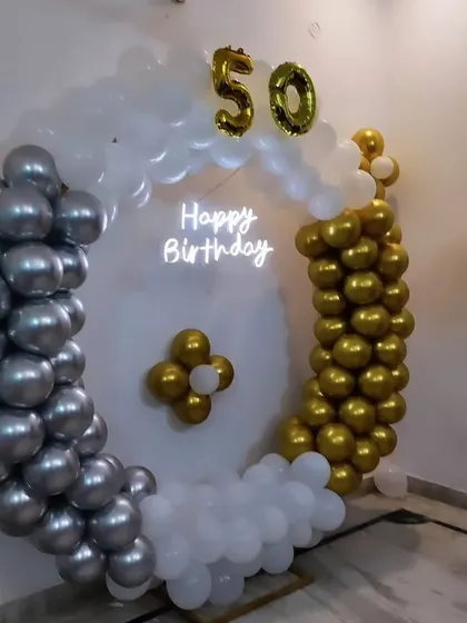 A 50th birthday decoration with a circular balloon frame. The setup uses silver, gold, and white balloons with a neon sign and the number '50' in foil balloons.