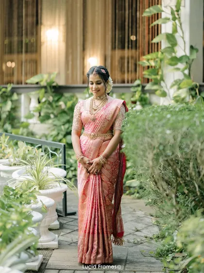 An elegant portrait of the bride in her pink silk saree, standing in a beautiful garden setting.