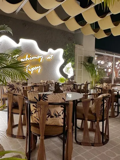 This seating area at Tipsy Town is defined by custom-designed chairs with unique wooden frames and printed fabric. The space is anchored by a neon sign and a wavy, suspended ceiling element, adding layers of texture and light.