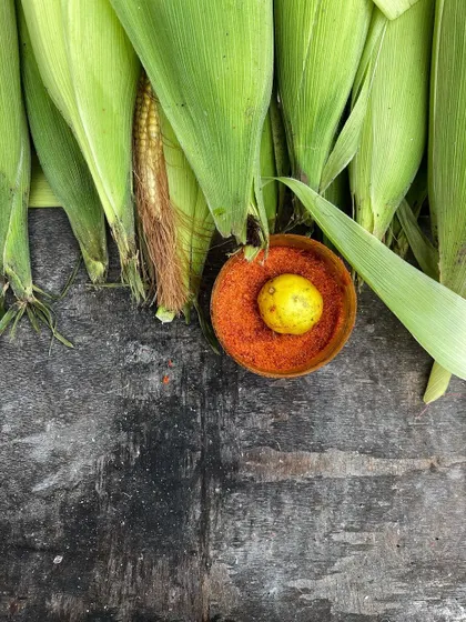 A simple, beautiful flatlay of fresh corn, with a lemon and a bowl of red chilli powder ready for seasoning.