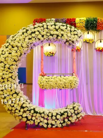 A stunning cradle ceremony setup featuring a crescent-shaped arch made of white flowers. The hanging cradle, also adorned with flowers, creates a beautiful focal point, while colorful lanterns add a festive touch.