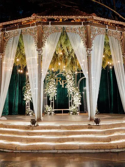 An elegant Sangeet stage with a deep green velvet backdrop, accented by modern gold arches and lush white floral arrangements. The couple's monogram adds a personalized touch to this sophisticated design.
