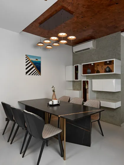 The dining room at Sobha Indraprastha features a modern black dining table and quilted chairs. Floating white shelves with internal lighting provide a stylish display area against a textured grey wall.