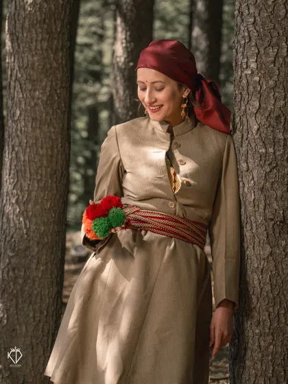 A beautiful solo portrait of the woman in her traditional Pahari dress, leaning against a pine tree. The details of her attire, like the colorful waist accessory, are highlighted.