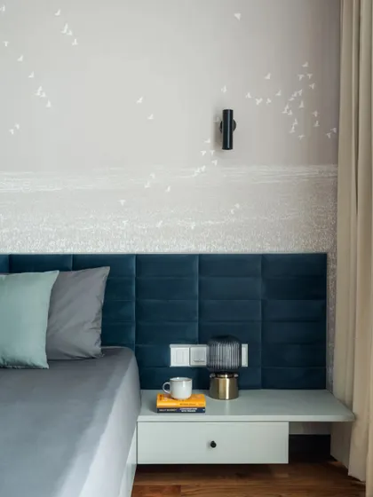 In this bedroom, a custom deep blue velvet headboard provides a touch of color and texture against a serene, hand-painted wallpaper depicting a distant shoreline. The floating nightstand and minimalist wall sconce maintain the room's clean lines, crafting a space that feels both elegant and deeply restful.