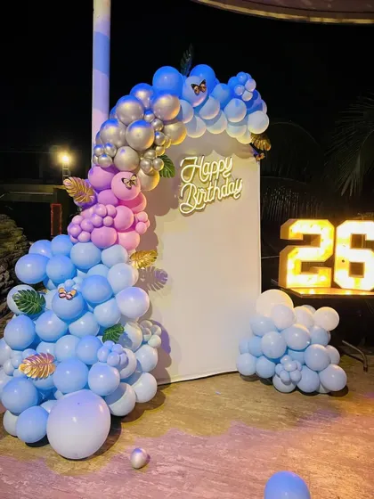 A beautiful outdoor birthday setup with a blue and pink theme. This decoration includes a balloon arch on a white backdrop and a light-up number '26'.