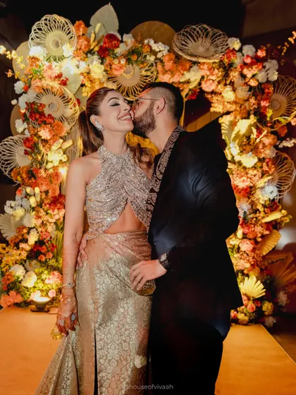 The couple shares an intimate moment against a stunning photo backdrop made of woven fans and an arch of autumn-toned florals, showcasing a unique and artistic installation.