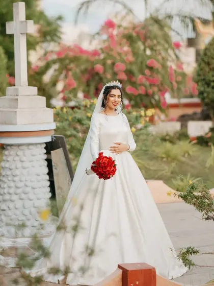 A stunning, regal portrait of a bride in her classic, long-sleeved satin gown, holding a bouquet of bold red roses.