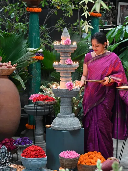 Preparing for Sankranthi celebrations at our store. A woman in a traditional saree lights a tiered stone temple lamp, surrounded by offerings of fresh flowers, embodying gratitude for the harvest season.