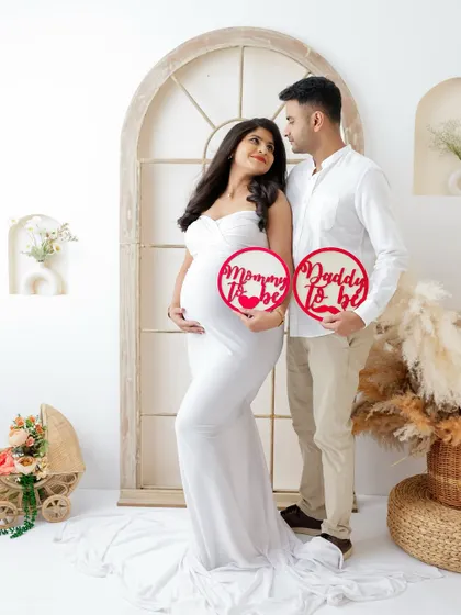 A classic and elegant couple's portrait. The mom-to-be in a white gown holds "Mommy to be" and "Daddy to be" signs with her partner.