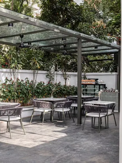 The outdoor seating area at Kana Cafe, covered by a glass canopy to provide a sun-kissed experience. The minimal furniture and concrete flooring enhance the raw, earthy, and breezy feel inspired by Bali.