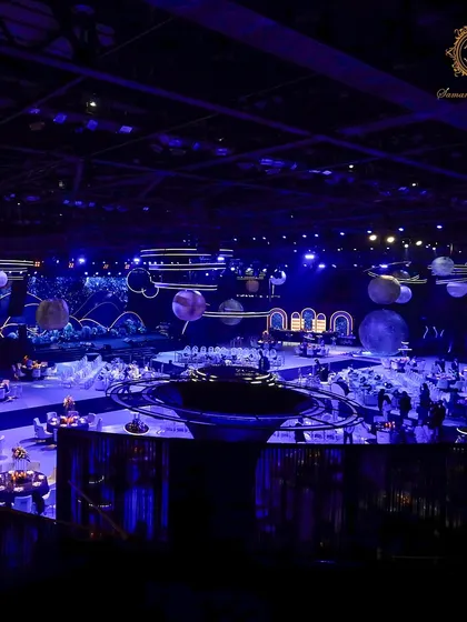 A wide shot capturing the full expanse of the cosmic-themed engagement party. The central bar, guest tables, and massive stage are all united under a deep blue, star-filled atmosphere.