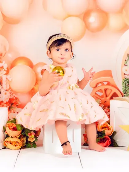 A baby girl sits elegantly in a peach-toned floral setup, holding a golden prop. She looks like a little queen on her throne.