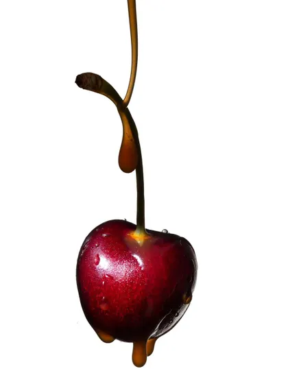 A single cherry with a delicate drip of caramel. The clean white background and focused lighting draw all attention to the interaction between the fruit's glossy skin and the sticky liquid.