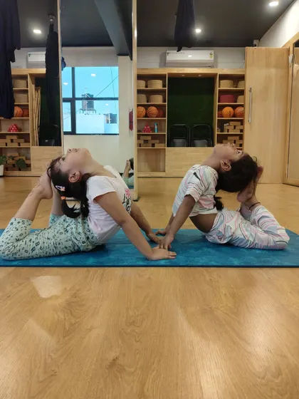 Two young yoginis practicing the One-Legged King Pigeon Pose together. Partner poses like this encourage friendship, trust, and mutual support on the mat.