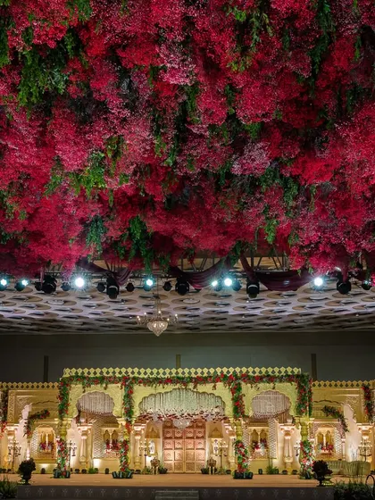 I transform grand ballrooms into royal courts. This wedding stage is defined by its magnificent ceiling, a dense canopy of crimson and green florals that creates an atmosphere of immersive, regal opulence and provides a breathtaking backdrop for the ceremony.