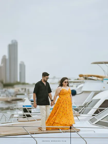 The couple holds hands while standing on the bow of a yacht, a classic pre-wedding pose that feels both adventurous and romantic against the city backdrop.