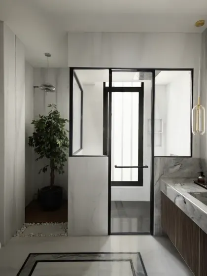 The son's bathroom in The House of Dancing Screens features a clean white and grey palette. A lighted mirror, chrome fittings, and an indoor plant add sophistication and a touch of nature to the space.