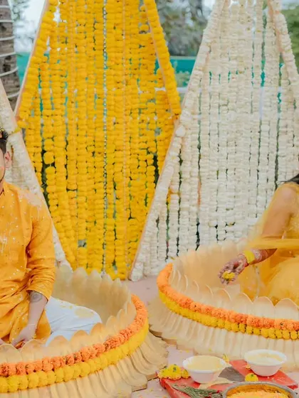 The bride and groom seated in traditional brass urlis decorated with marigolds for their Haldi ceremony. The backdrop features cone-shaped structures covered in yellow and white flowers, a unique design element for a memorable setup.