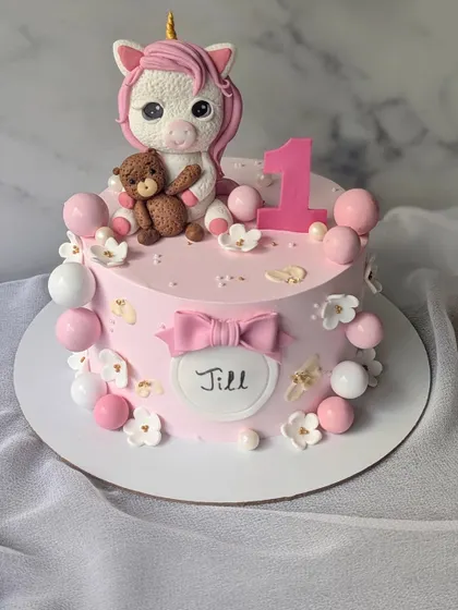 Unleashing magic with this adorable unicorn cake for a first birthday. It features a cute, handcrafted unicorn holding a teddy bear, surrounded by pink and white spheres.