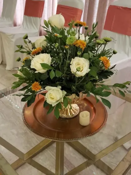 A classic centerpiece for a reception table, featuring white roses and orange flowers in a golden vase, placed on a copper charger plate.