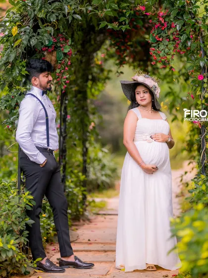 A classic portrait of an expecting mother in a simple white gown and a stylish hat. Her partner looks on from the background, framed by a beautiful floral archway in a lush Bangalore garden.