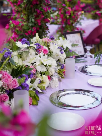 A close-up of the tablescape, featuring a lush floral centerpiece, silver charger plates, and a soft lavender tablecloth. The out-of-focus bougainvillea adds a beautiful pop of color.